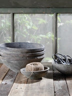A rustic collection of dark gray ceramic bowls stacked on a weathered wooden table, with a ball of twine in a small bowl and a pair of scissors in a larger bowl, set against a soft-focus window view of green foliage.