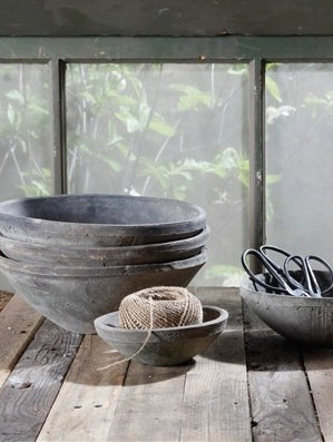 A rustic collection of dark gray ceramic bowls stacked on a weathered wooden table, with a ball of twine in a small bowl and a pair of scissors in a larger bowl, set against a soft-focus window view of green foliage.
