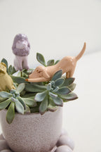 A close-up of a speckled light pink ceramic planter filled with green succulent plants and small pastel ceramic dog figurines, set against a clean white background.