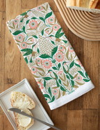 The Hummingbird Flour Sack Tea Towel, featuring pink flowers and green leaves, is displayed on a wooden table beside a breadboard with a loaf of bread and a plate holding a buttered slice and butter knife.