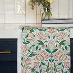 The Hummingbird Flour Sack Tea Towel, featuring a vibrant floral and bird design, hangs over a dark blue kitchen drawer. In the background, a glass vase with flowers and two stacked books sit on the countertop.