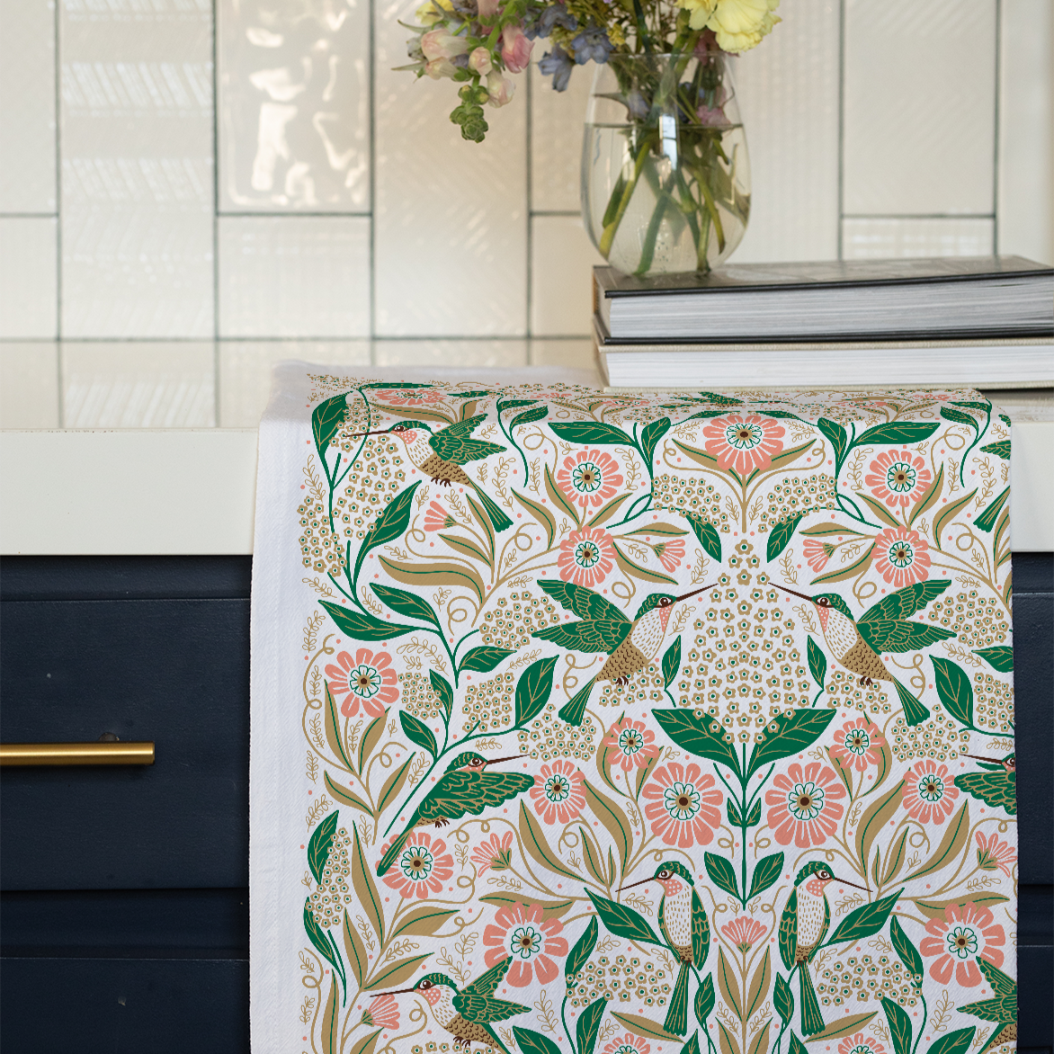 The Hummingbird Flour Sack Tea Towel, featuring a vibrant floral and bird design, hangs over a dark blue kitchen drawer. In the background, a glass vase with flowers and two stacked books sit on the countertop.