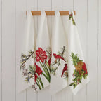 Four Christmas Bloom Kitchen Towels featuring festive birds, red flowers, and pinecones hang side by side on a wooden rack against a white paneled wall.