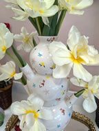 The Ditsy Floral Tulipiere Vase, featuring colorful floral patterns, displays large pale yellow flowers on a gold tray near potted plants, set against a soft pink wall.