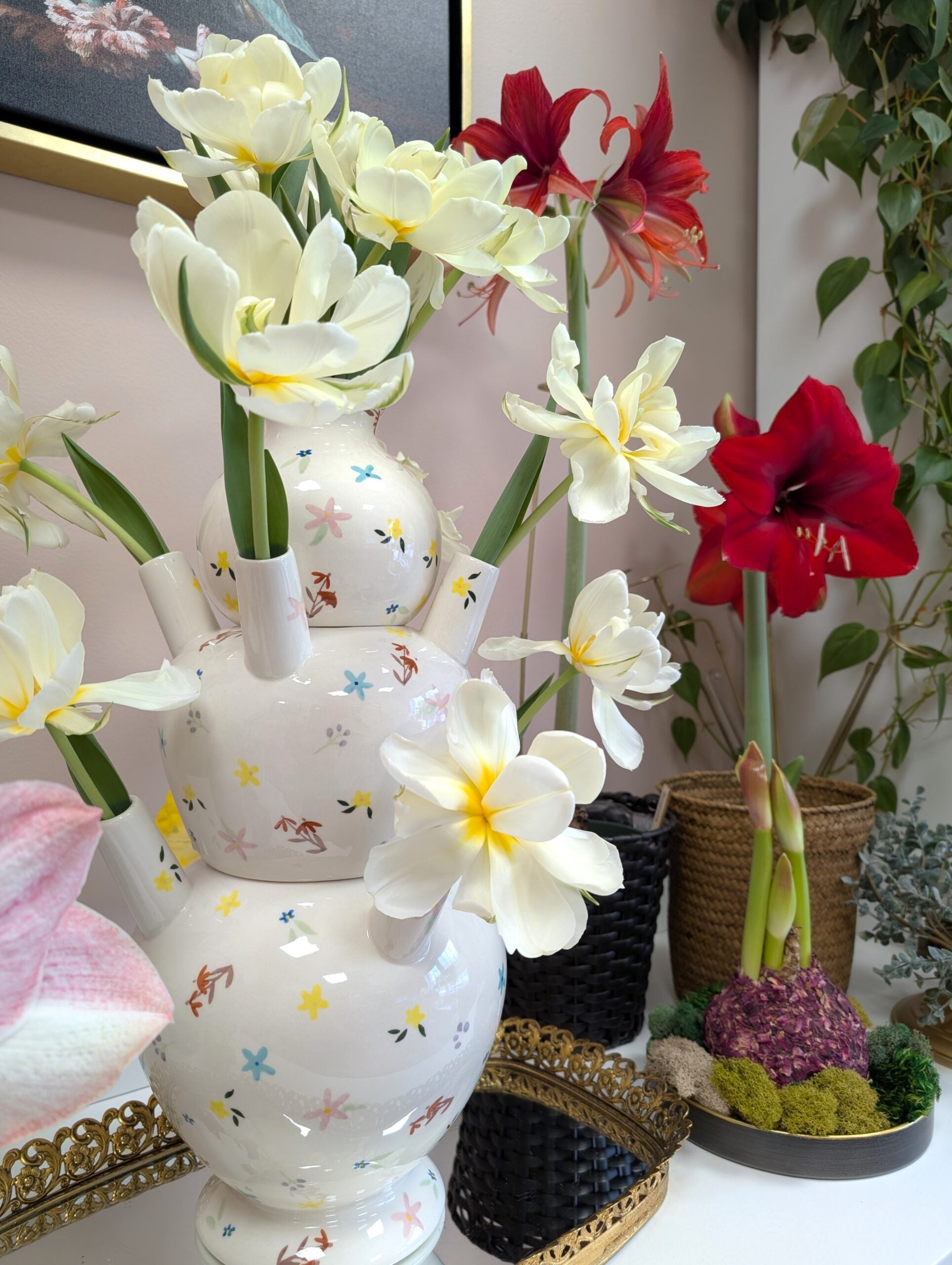 The Ditsy Floral Tulipiere Vase, adorned with colorful patterns, holds white and yellow flowers on a table beside red amaryllis in woven pots, a decorative tray, and plants set against a pale pink wall with a leafy vine.