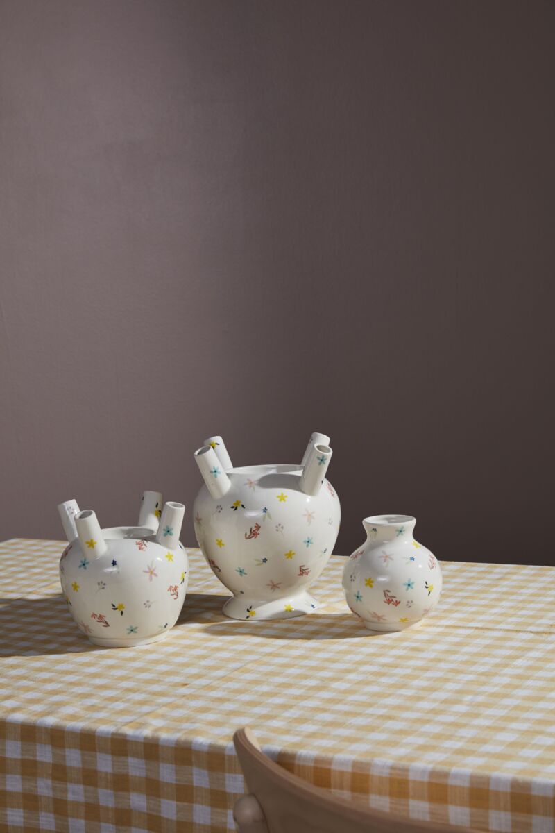 Three white ceramic vases, featuring the Ditsy Floral Tulipiere Vase with colorful floral patterns, are displayed on a yellow and white checkered tablecloth in front of a plain mauve wall. One vase is heart-shaped; two are round with tube-like openings.