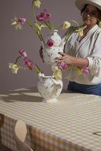 A woman in a hat arranges pink and yellow tulips in two Ditsy Floral Tulipiere Vases on a table with a yellow and white checkered tablecloth.