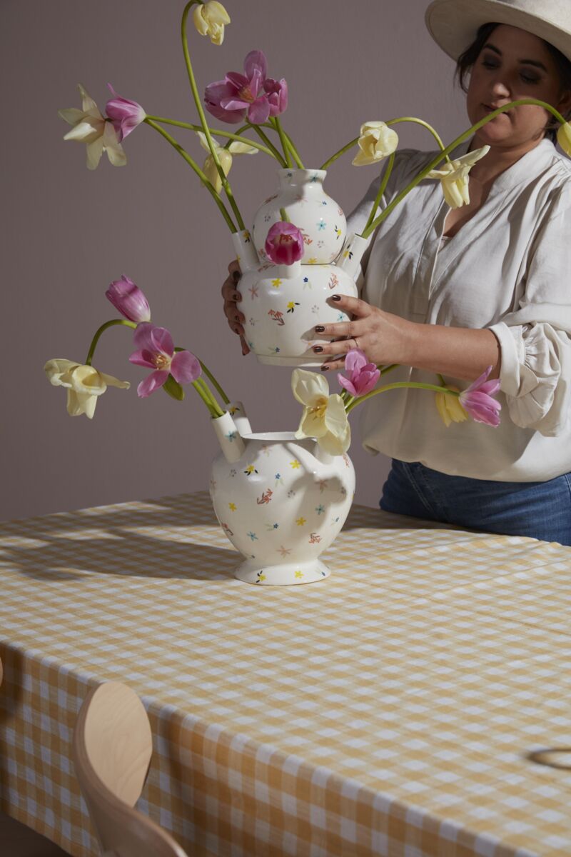 A woman in a hat arranges pink and yellow tulips in two Ditsy Floral Tulipiere Vases on a table with a yellow and white checkered tablecloth.