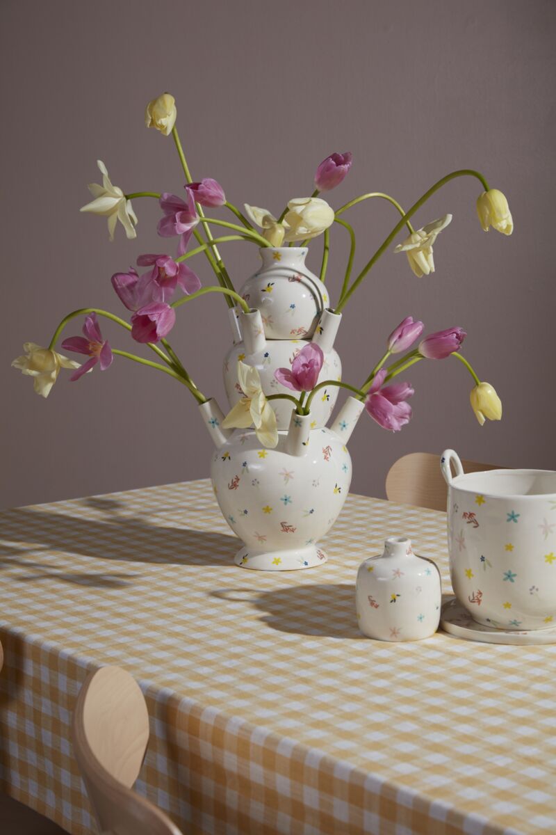 The Ditsy Floral Tulipiere Vase holds pink and yellow tulips on a checkered tablecloth, while a matching jug and sugar bowl rest nearby beside light wooden chairs.