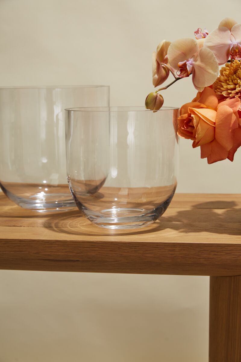Two Fat Bowl Paperwhite Forcing Vases - Medium rest on a wooden table; one displays orange roses and light pink orchids, while the other is empty. A pale beige wall in the background completes the elegant scene.