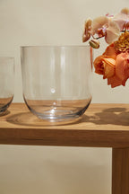 The Fat Bowl Paperwhite Forcing Vase - Large, made of clear glass, sits on a wooden table partially filled with orange roses and light pink orchids. The background is a plain, light beige color.