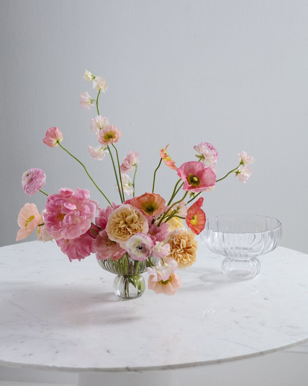 A pastel floral arrangement in a clear glass vase on a white marble table, with a clear glass pedestal bowl beside it against a light gray background.
