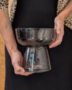 A person holding a two-tiered transparent smoky glass bowl stand against a black dress, showing the clear glass structure.
