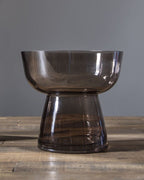 A smoky translucent glass pedestal bowl with a wide rim and flared base, shown on a wooden tabletop against a plain gray wall. 