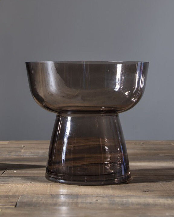 A smoky translucent glass pedestal bowl with a wide rim and flared base, shown on a wooden tabletop against a plain gray wall. 