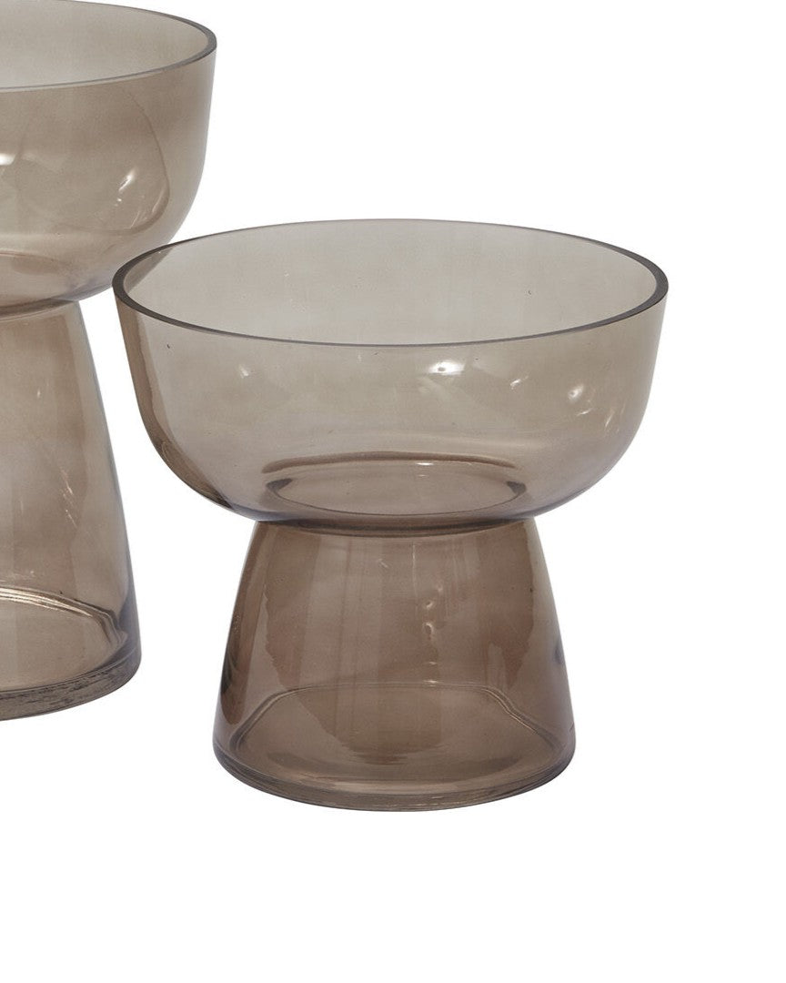 A translucent smoky brown glass pedestal bowl with a wide rim and tapered base, shown on a plain white background with another similar bowl partially visible behind it.