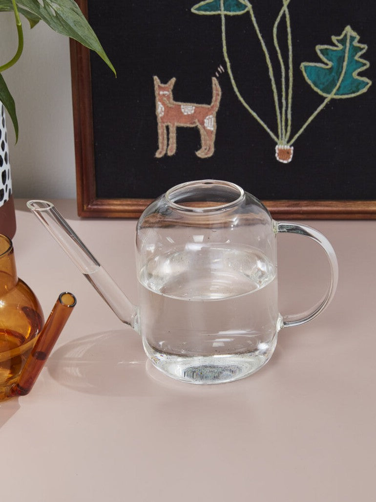 A clear glass watering can filled with water, placed on a light pink tabletop. A dark framed wall hanging with a stylized dog and plant is behind it, and part of an amber glass container and green leaves are visible at the edges.