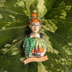 Bulb & Blossom's Plant Lady Glass Ornament features a red-haired woman with a bun, glasses, and green headband holding leafy plants with glitter accents. A ribbon below labels her an official “PLANT LADY".
