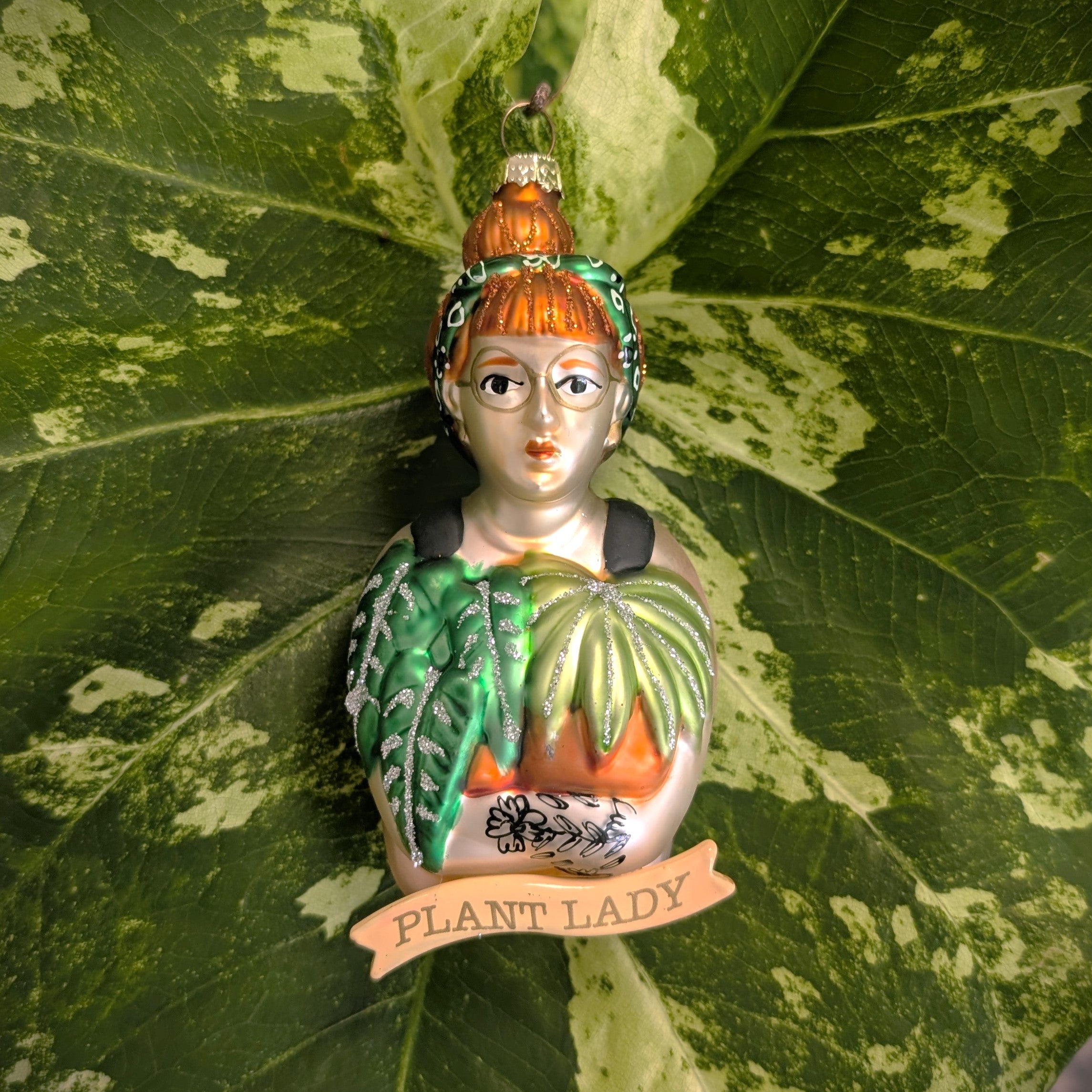 Bulb & Blossom's Plant Lady Glass Ornament features a red-haired woman with a bun, glasses, and green headband holding leafy plants with glitter accents. A ribbon below labels her an official “PLANT LADY".