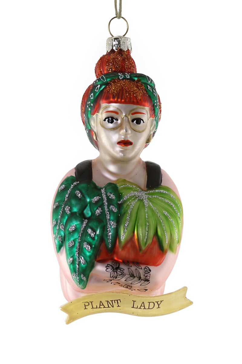 Bulb & Blossom's Plant Lady Glass Ornament features a red-haired woman with a bun, glasses, and green headband holding leafy plants with glitter accents. A ribbon below labels her an official “PLANT LADY".