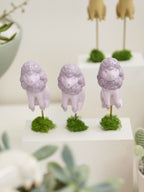 Three small lavender poodle figurines standing on mossy bases, displayed on white pedestals with blurred plants in the foreground.