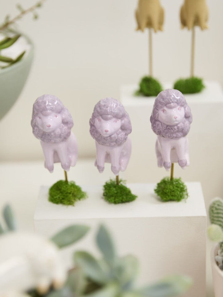 Three small lavender poodle figurines standing on mossy bases, displayed on white pedestals with blurred plants in the foreground.