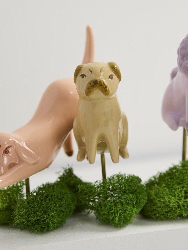 A ceramic dog figurine in beige and brown stands on a green moss base, flanked by partial figurines in peach and lavender, against a clean white background.