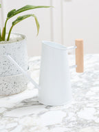 A white metal watering can with a light wood handle, shown on a marble countertop next to a potted plant.
