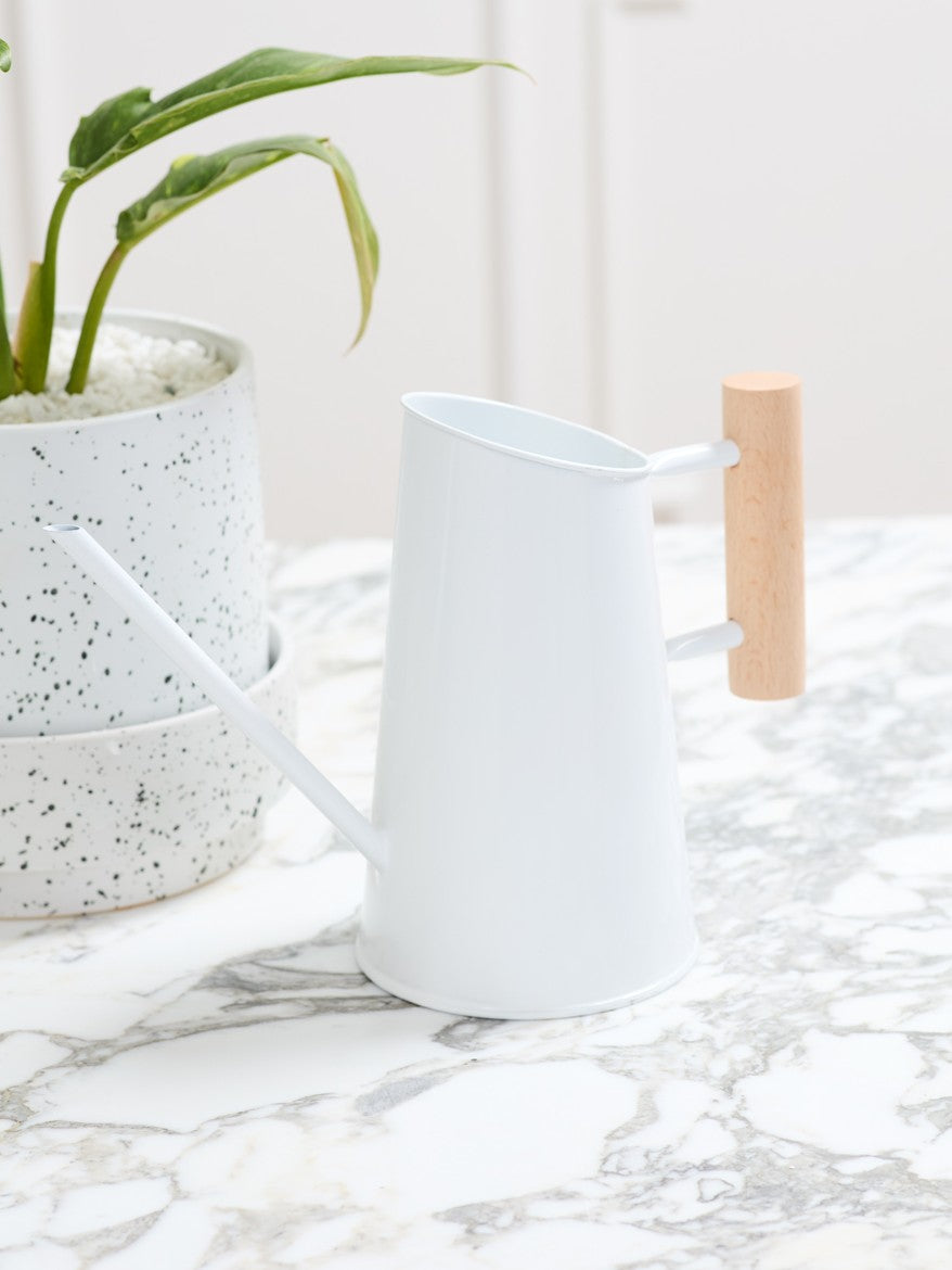 A white metal watering can with a light wood handle, shown on a marble countertop next to a potted plant.