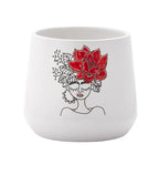 The Red Flower Crown Ceramic Pot is a white ceramic planter featuring a minimalist woman's face with closed eyes and a bold red flower with leafy accents as her hair.