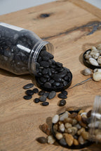 Black stones spilling out of a transparent container on a wooden surface with other types of stones.