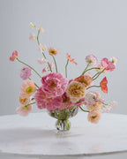A pastel floral arrangement in a clear glass vase on a white marble table against a light gray background. The bouquet features pink, peach, cream, and white flowers with green stems.