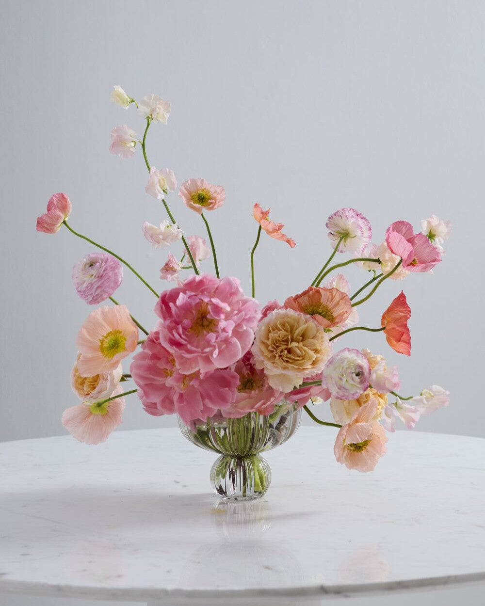 A pastel floral arrangement in a clear glass vase on a white marble table against a light gray background. The bouquet features pink, peach, cream, and white flowers with green stems.