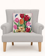 A square decorative throw pillow featuring a colorful floral tapestry design of red, pink, and orange tulips with green leaves and small blue butterflies on a cream background, displayed on a light gray upholstered armchair.