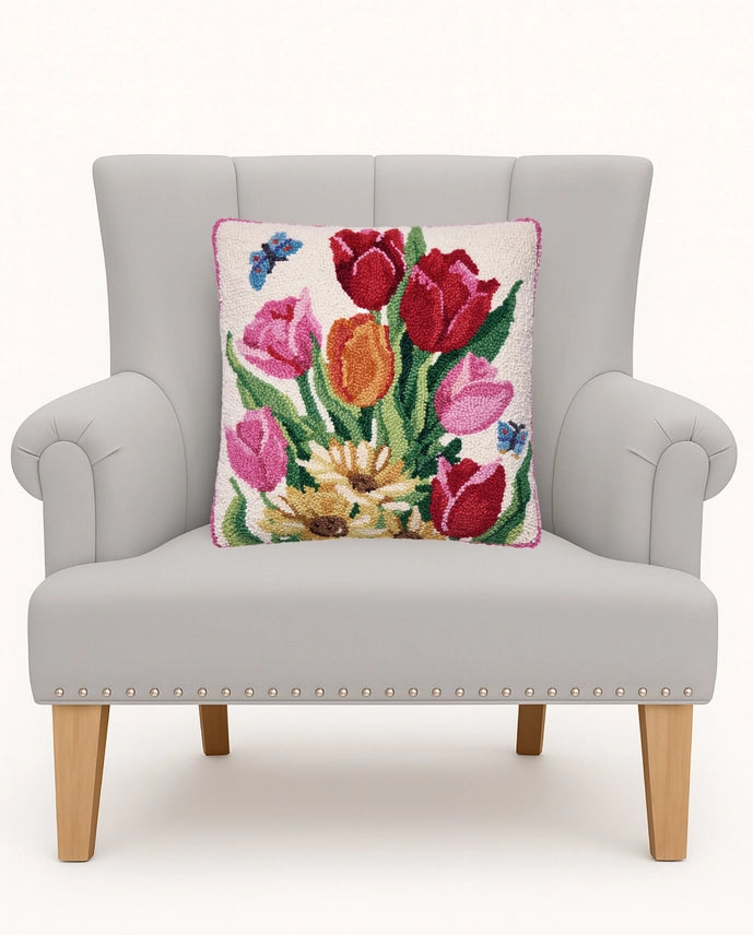 A square decorative throw pillow featuring a colorful floral tapestry design of red, pink, and orange tulips with green leaves and small blue butterflies on a cream background, displayed on a light gray upholstered armchair.