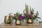 Three Tulum Pots are on a wooden table before a white brick wall; two hold assorted purple, pink, and green flowers and foliage, while the third pot is empty.