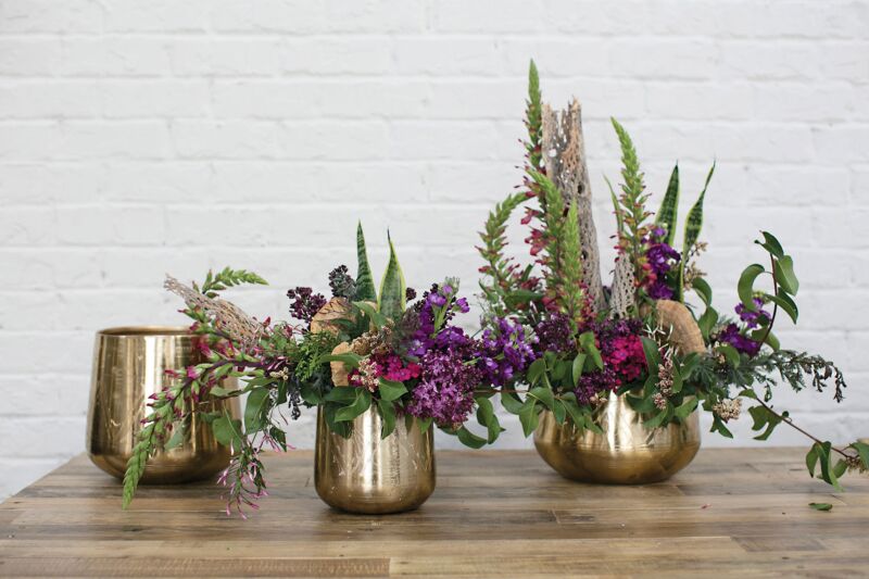 Three Tulum Pots are on a wooden table before a white brick wall; two hold assorted purple, pink, and green flowers and foliage, while the third pot is empty.